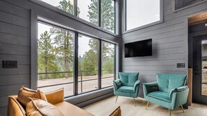 Ein modernes Wohnzimmer mit großen Fenstern, einem Sofa, zwei Sesseln und einem an der Wand montierten Flachbildfernseher.
