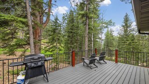 A deck with a barbecue grill and two chairs, surrounded by tall trees. - Modern Black Hills Cabin · Romantic Getaway · Hot Tub · Near Deadwood (Sturgis)