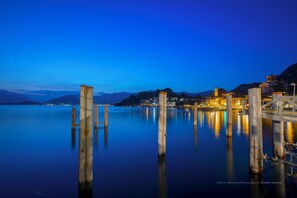 A calm waterfront at dusk with wooden posts in the water, a bridge, and buildings with lights in the background. - Bucaneve Apartment (Casalzuigno)