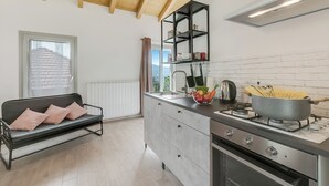 Une cuisine moderne équipée d’une cuisinière à gaz, d’un évier et d’une étagère noire placée au-dessus. On y trouve une fenêtre avec des stores et un radiateur blanc fixé au mur.