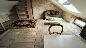 A loft bedroom with a sloped ceiling, a wooden staircase, a green sofa with cushions, a small table with a floral tablecloth, and a patterned rug.