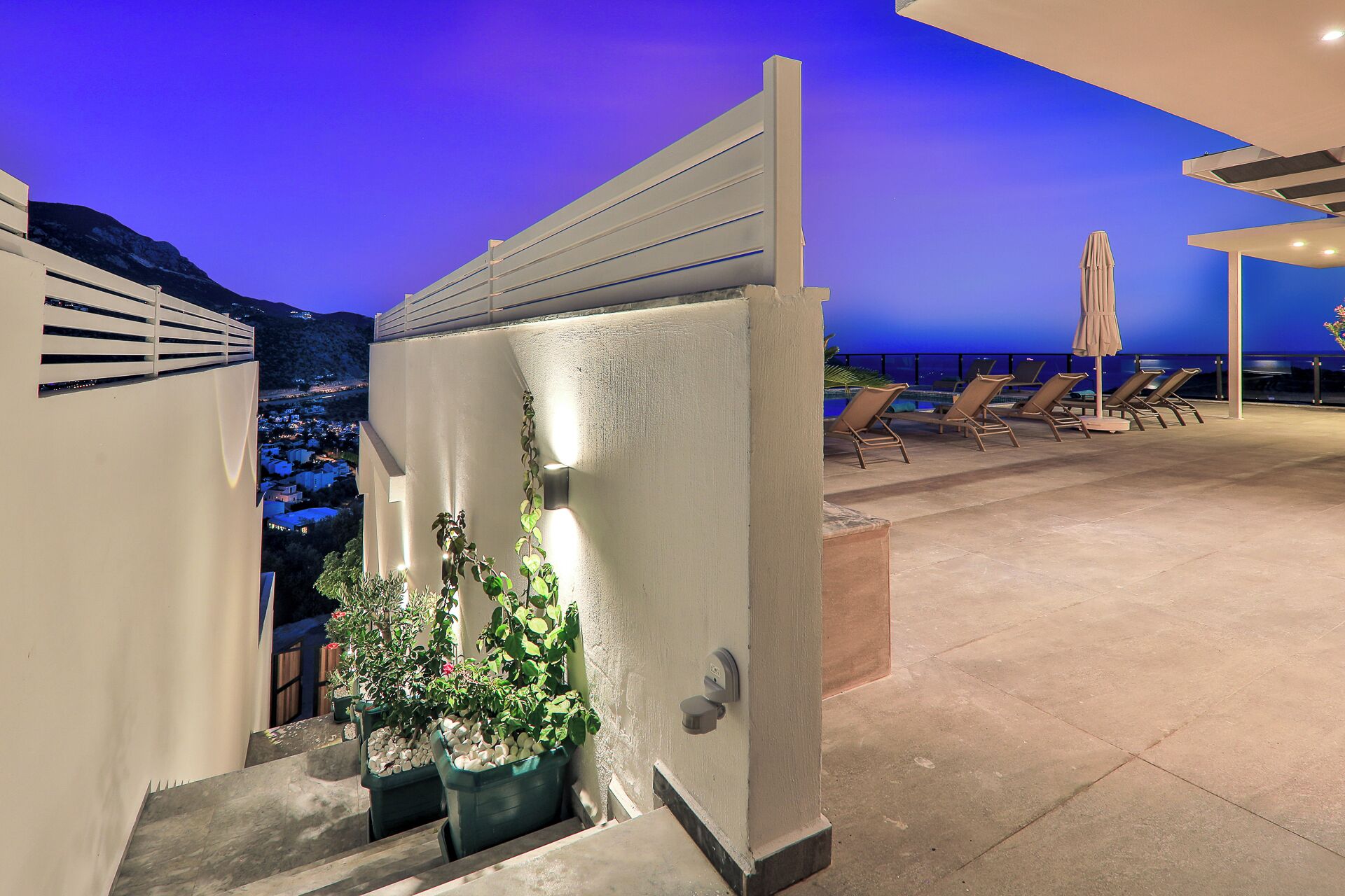 Terraza en la azotea con vistas a la ciudad al atardecer, plantas en macetas y tumbonas.
