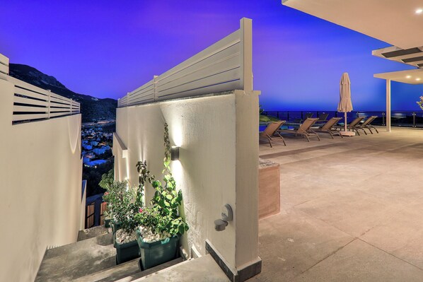 A rooftop terrace with a view of the city at dusk, potted plants, and sun loungers.
