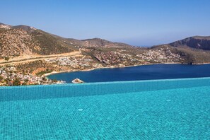 A coastal town with a clear blue sea, a swimming pool, and mountainous terrain in the background.
