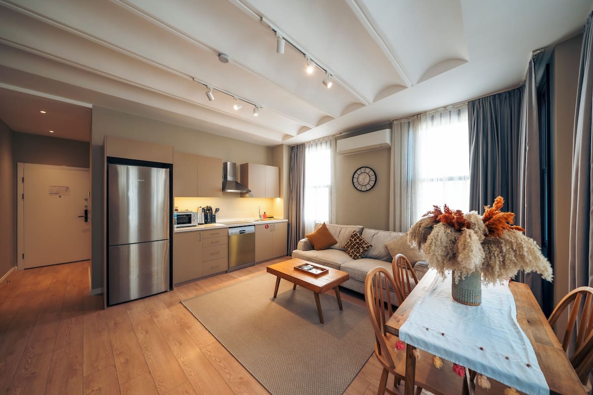 A modern kitchen with stainless steel appliances, a dining area with a wooden table and chairs, and a living room with a sofa and a decorative vase.