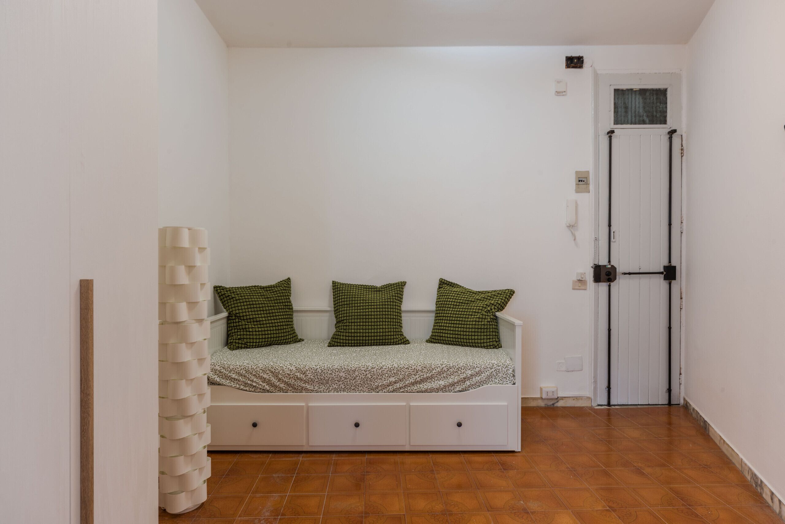 Une petite chambre avec un lit simple, un mur blanc, une porte en bois et un sol carrelé.