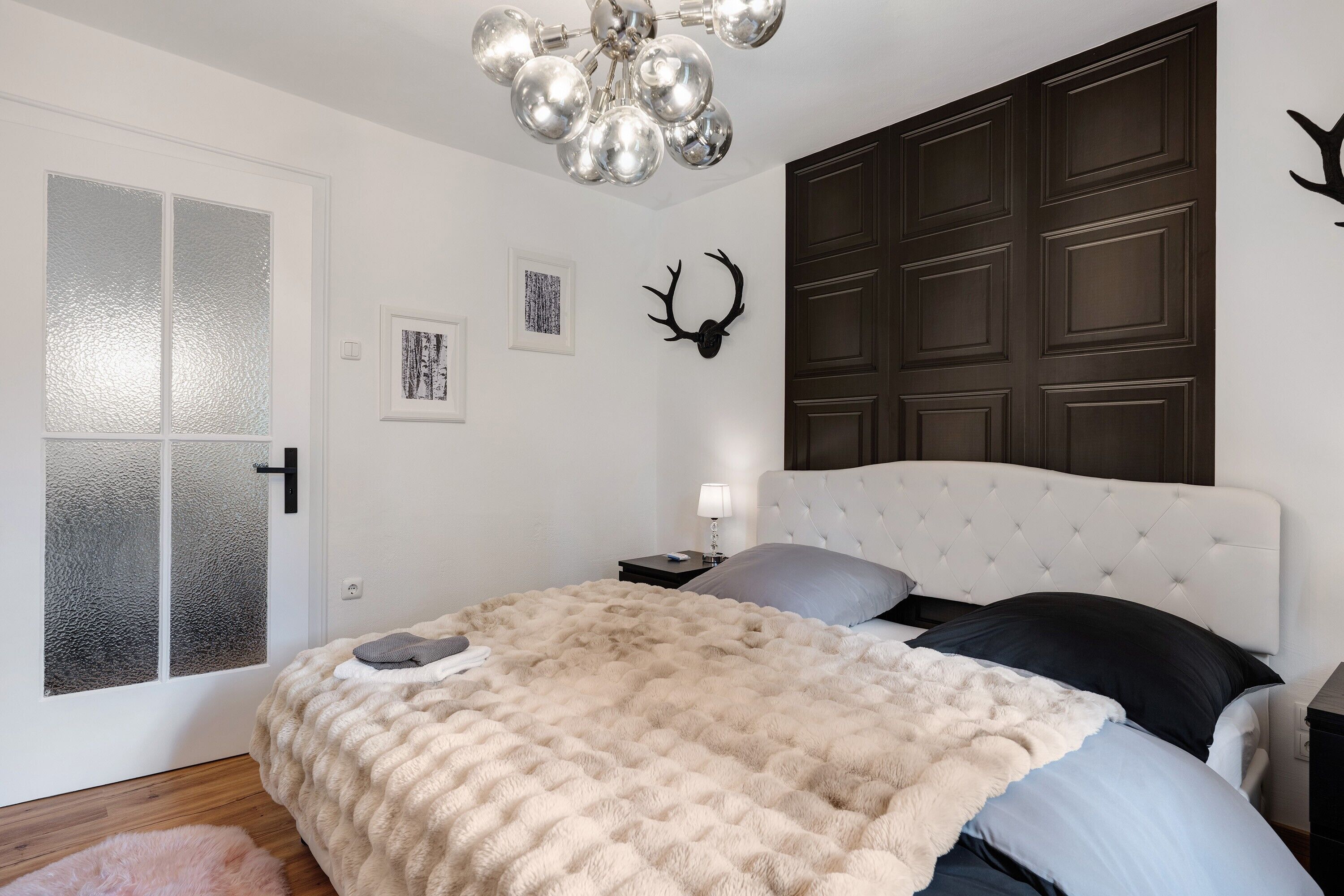 Une chambre avec une tête de lit capitonnée, une couverture beige moelleuse, un plancher en bois, une porte blanche dotée de panneaux en verre dépoli et une décoration murale représentant une tête de cerf.