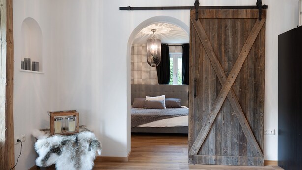 Een kamer met een houten schuifdeur, een bed met een grijze hoofdbord, een kroonluchter en een klein tafeltje met een bontmantel.