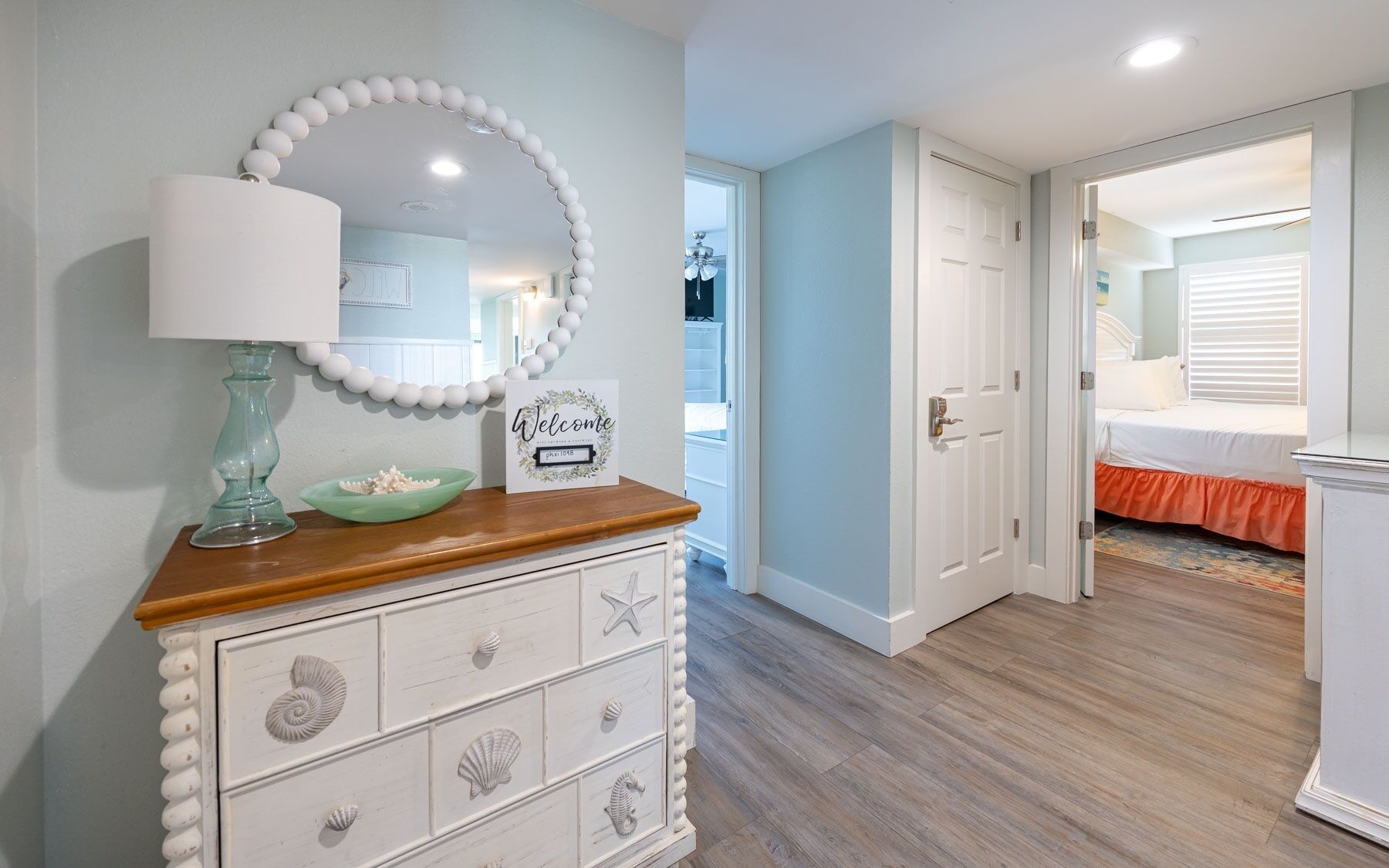 A room with a wooden dresser, a mirror, a welcome sign, and a bed with an orange skirt.