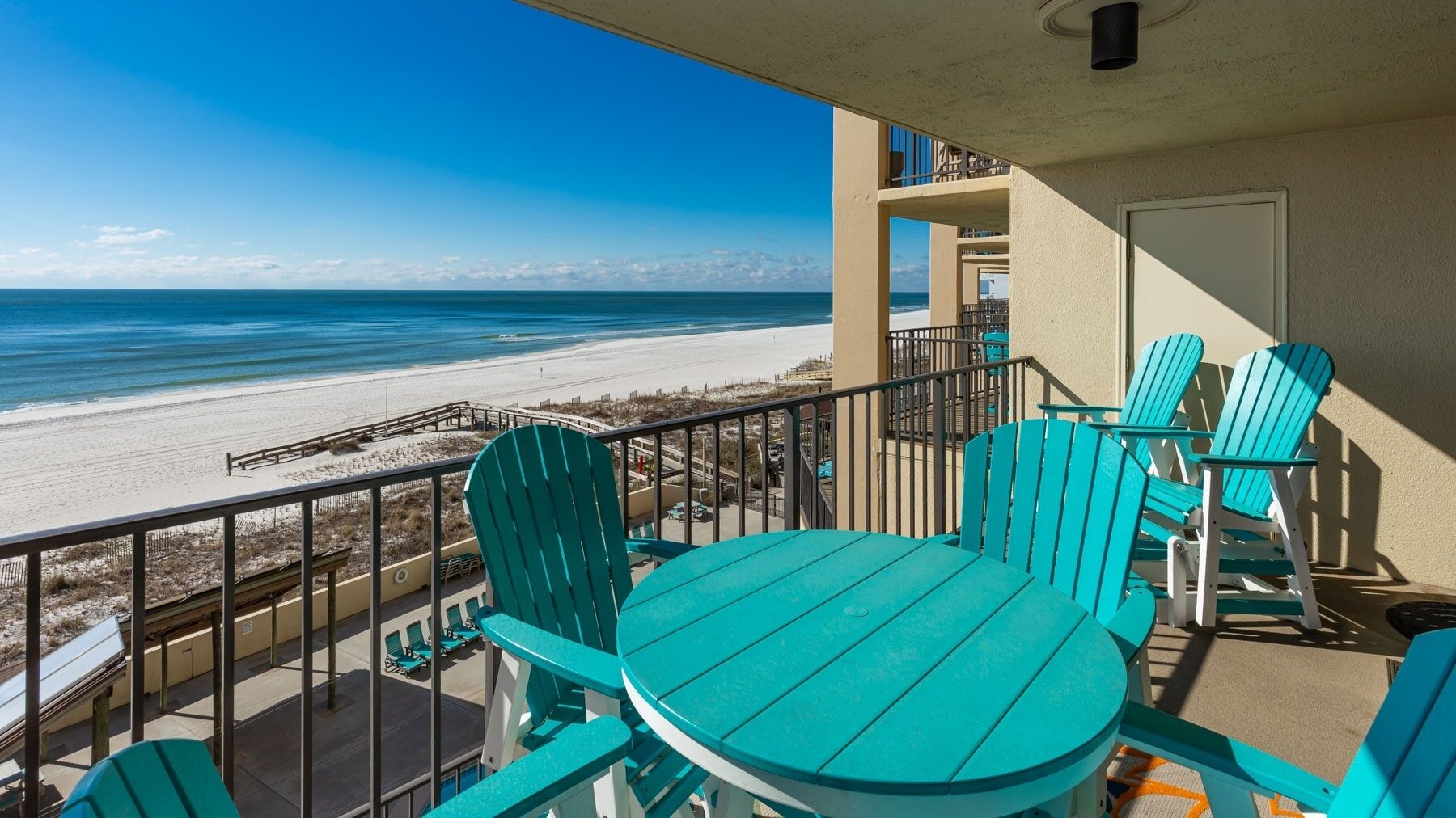 A balcony with turquoise chairs and a table overlooking a beach.