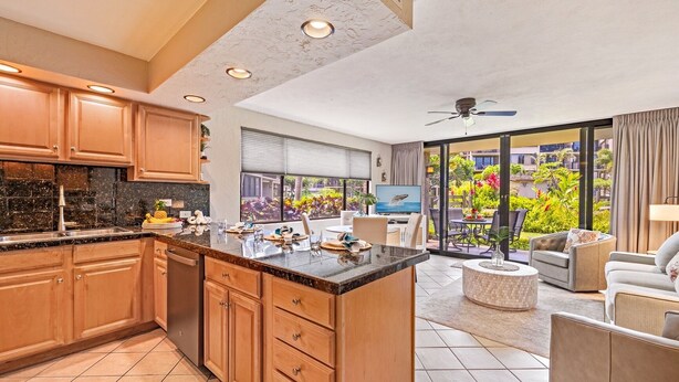 A modern kitchen with wooden cabinets and a large island, a dining area with a TV, and a balcony with a view of a lush garden. - Papakea G101 1bed/1bath New Remodel 1 Bedroom Condo (Lahaina)