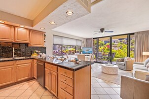 Una cocina moderna con armarios de madera y una isla grande, un comedor con televisor, y un balcón con vistas a un jardín exuberante.