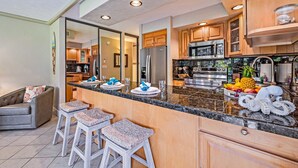 A modern kitchen with granite countertops, stainless steel appliances, and wooden cabinets. There are three stools at the counter and a refrigerator in the background. - Papakea G101 1bed/1bath New Remodel 1 Bedroom Condo (Lahaina)