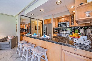Una cocina moderna con encimeras de granito, electrodomésticos de acero inoxidable y armarios de madera. Hay tres taburetes en la encimera y un refrigerador al fondo.