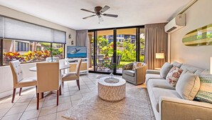 A modern living room with a television, seating area, and a view of a lush garden. - Papakea G101 1bed/1bath New Remodel 1 Bedroom Condo (Lahaina)