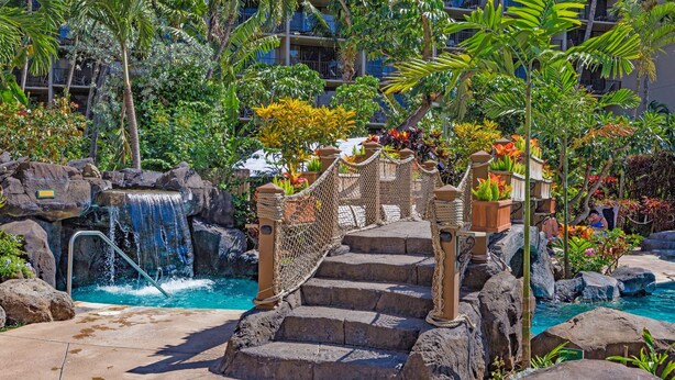 A pool area with a waterfall, stone steps, and tropical plants. - Kaanapali Shores 432 Studio Condo Condo (lahaina)