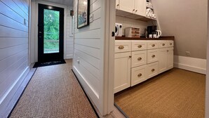 A narrow hallway with a door leading to a balcony, a kitchen area with white cabinets and a microwave, and a framed picture on the wall. - Killington A-frame 2 Bedroom Home (Killington)