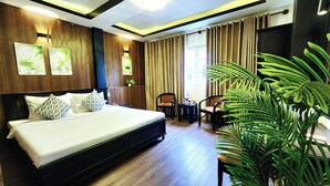 A hotel room with a large bed, wooden panel walls, a small table with chairs, and a large potted plant. - Rosa May Hotel (Ho Chi Minh City)