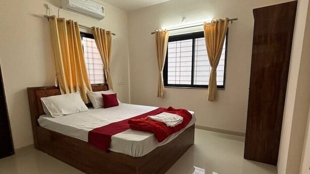 A bedroom with a wooden bed, white bedding, red and white pillows, and beige curtains. - 1BHK Comfy Stay 5 (Pune)