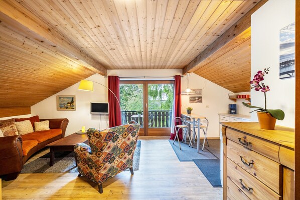 Une pièce chaleureuse avec un plafond en bois, un canapé, un fauteuil à motifs, une table à manger et un coin cuisine.
