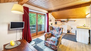 Ein gemütliches Zimmer mit Holzboden, einem Fernseher, einem gemusterten Sessel, einem kleinen Tisch mit Kerzen und einer Glastür zu einem Außenbereich.