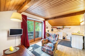 Et hyggeligt rum med trælofter, et fjernsyn, en polstret stol med mønster, et lille bord med stearinlys og en skydedør af glas, der fører til et udendørs område.