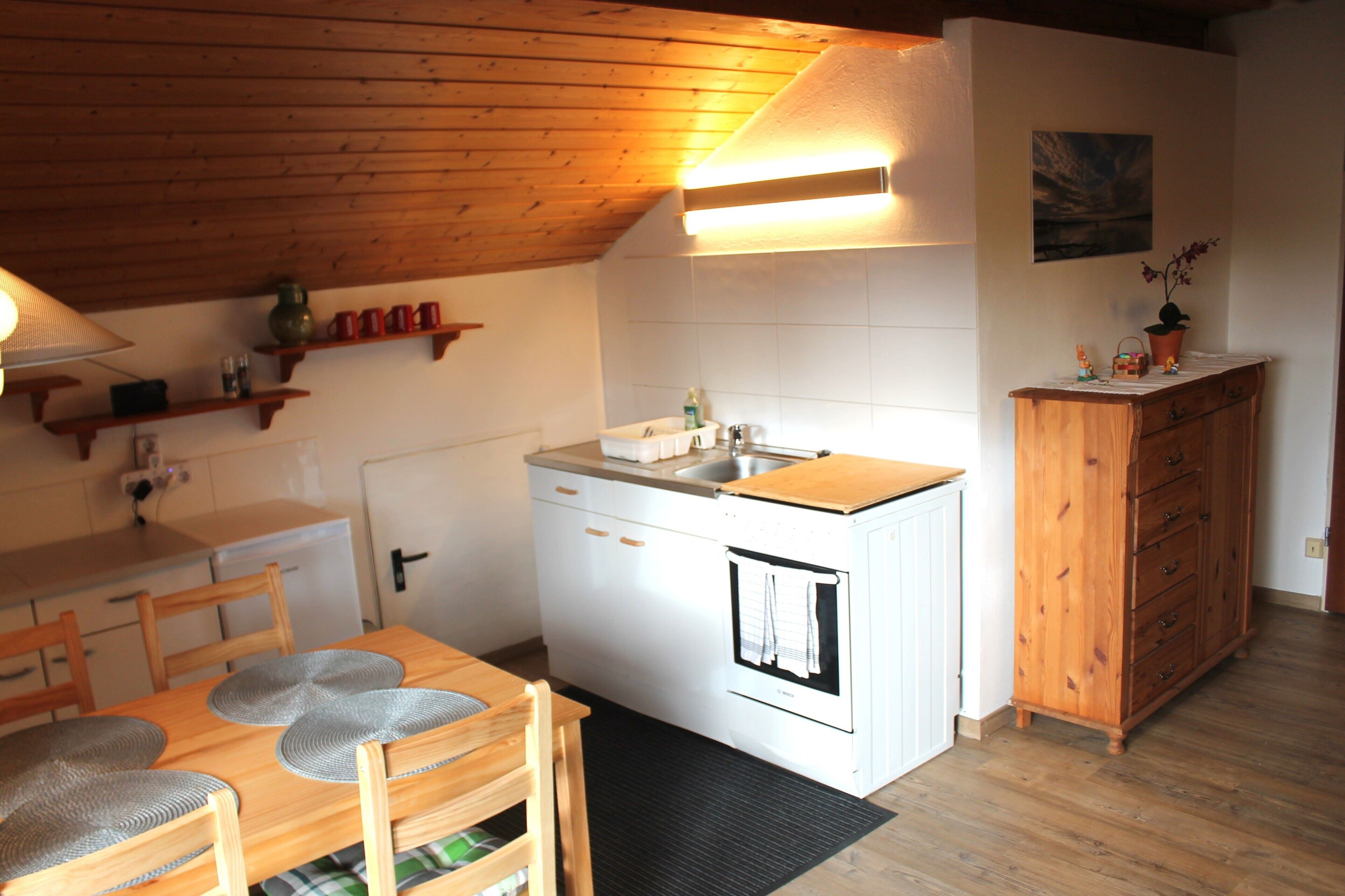 A compact kitchen with a wooden ceiling, a white refrigerator, a wooden cabinet, and a dining table with chairs.