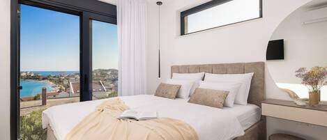 A modern bedroom with a large bed, a desk, and a view of the sea.