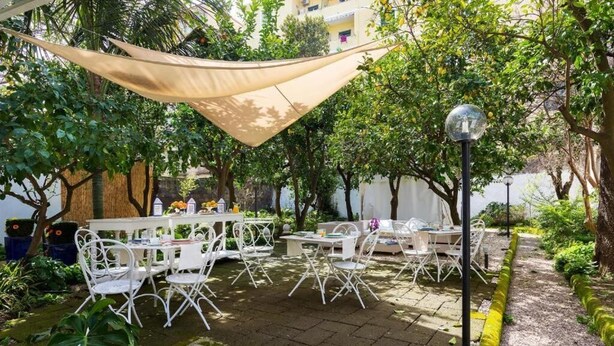 An outdoor patio area with white metal tables and chairs, surrounded by greenery and a yellow-painted curb. - Scaramellino Accommodation (Vico Equense)