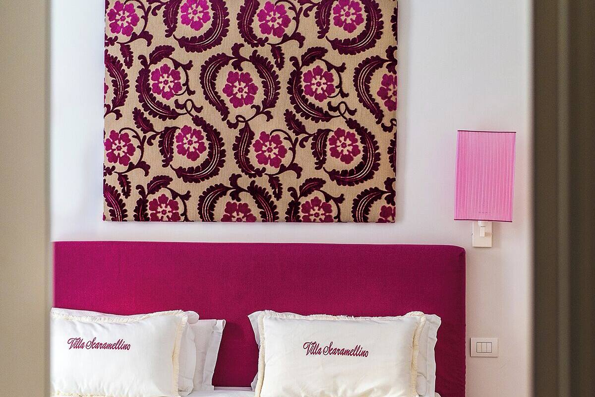 Un dormitorio con una cama, dos almohadas, una silla roja, una lámpara rosa y un tapiz con diseño en la pared.