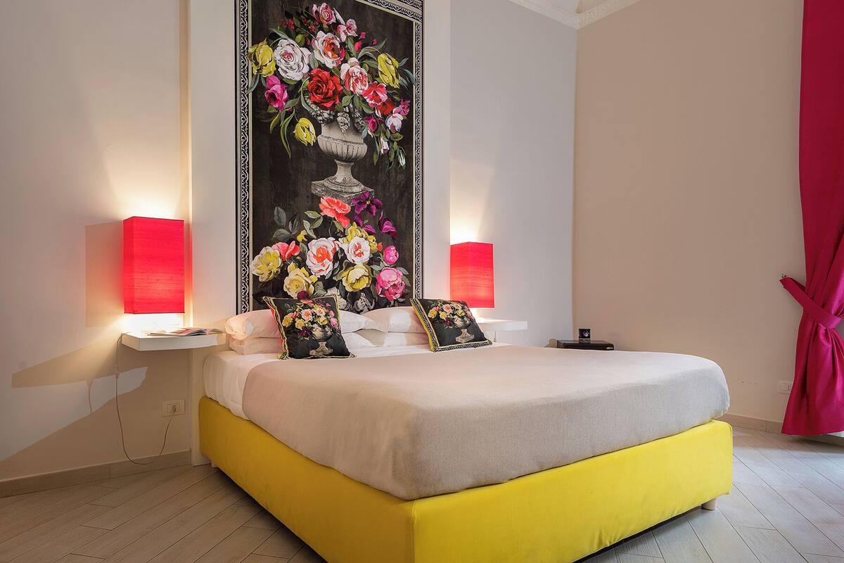 Un dormitorio con una cama, dos lámparas de noche, un cuadro grande con motivos florales y una cortina roja.