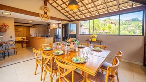 A spacious dining area with a large wooden table and chairs, a kitchen in the background, and a view of greenery outside. - Bz25 Casa com Pisc Geribá 250m da Praia (Buzios)