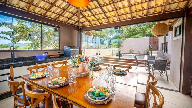 A dining area with a wooden table set for a meal, large windows, and a view of the outdoors. - Bz25 Casa com Pisc Geribá 250m da Praia (Buzios)