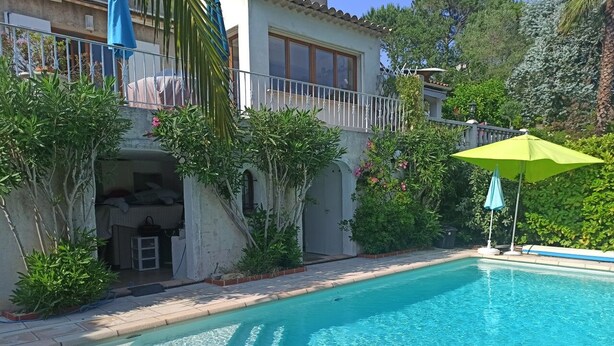 Ein Schwimmbecken mit einem grünen Sonnenschirm, umgeben von üppiger Bepflanzung und einem Gebäude mit Balkon.
