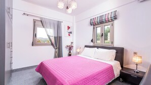 A bedroom with a bed, bedside tables, a window with curtains, a chandelier, and a view of the outdoors. - Villa Klio (Pitsidia Heraklion Crete)