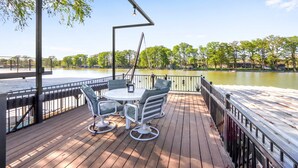 Een houten terras met een tafel en stoelen, uitkijkend op een rustig wateroppervlak en een rij bomen.