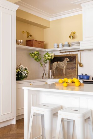 Una cocina con armarios blancos, una isla central con taburetes, un cuadro de un perro y un jarrón con flores.