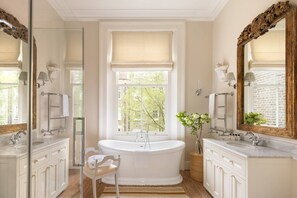 A bathroom with a freestanding bathtub, two sinks, a large mirror, and a window with a view of greenery.