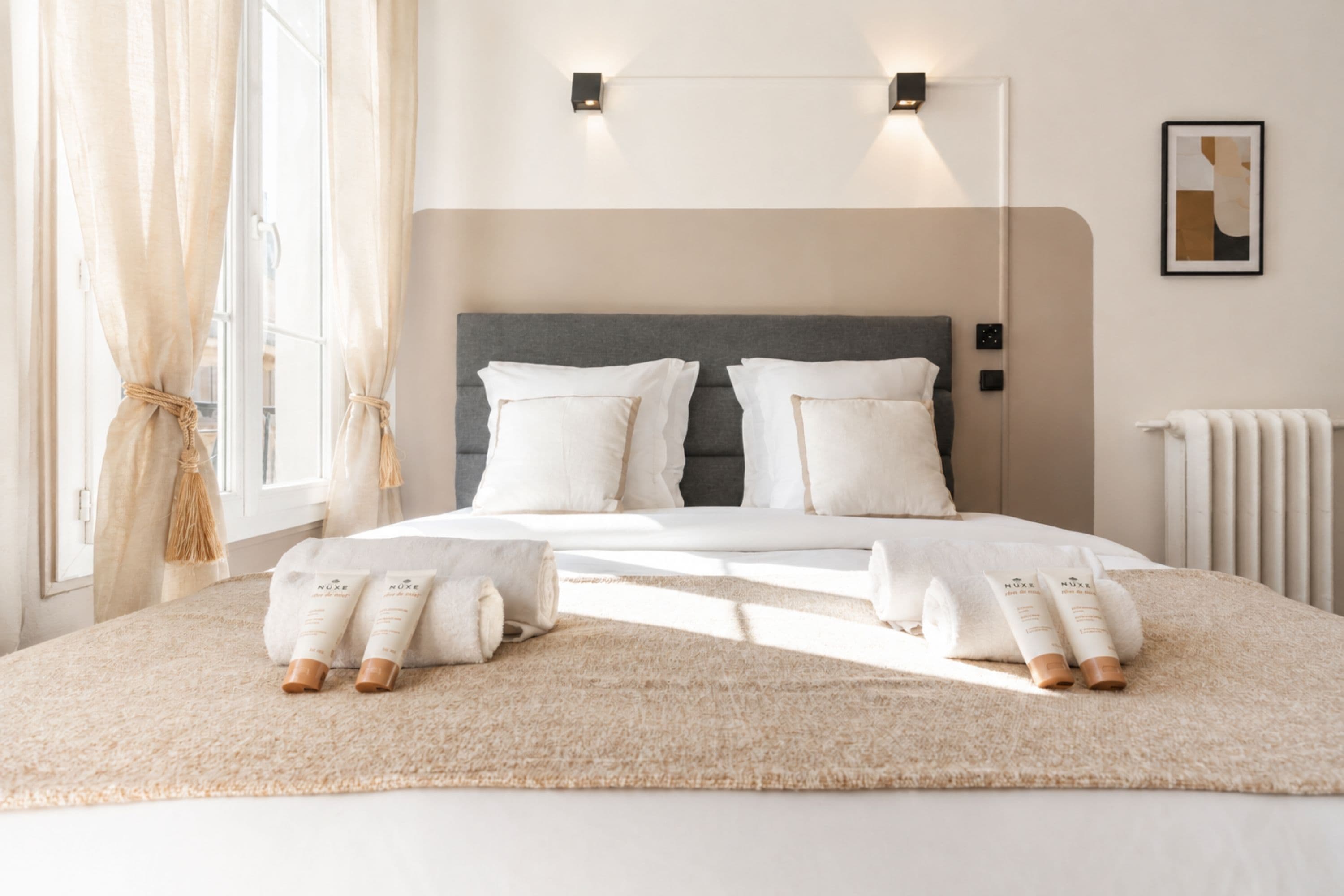 A neatly made bed with pillows and rolled towels, a bedside table, a wall-mounted light fixture, and a radiator.