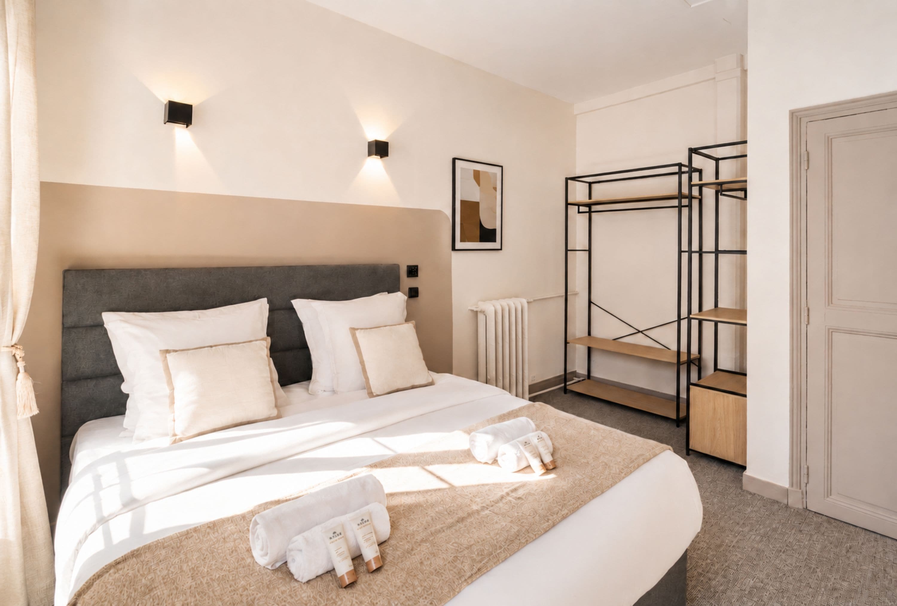 A hotel room with a bed, a radiator, a wall-mounted shelf, and a door.