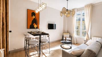 A modern living room with a sofa, dining area with chairs, a TV mounted on the wall, and a large window with curtains.
