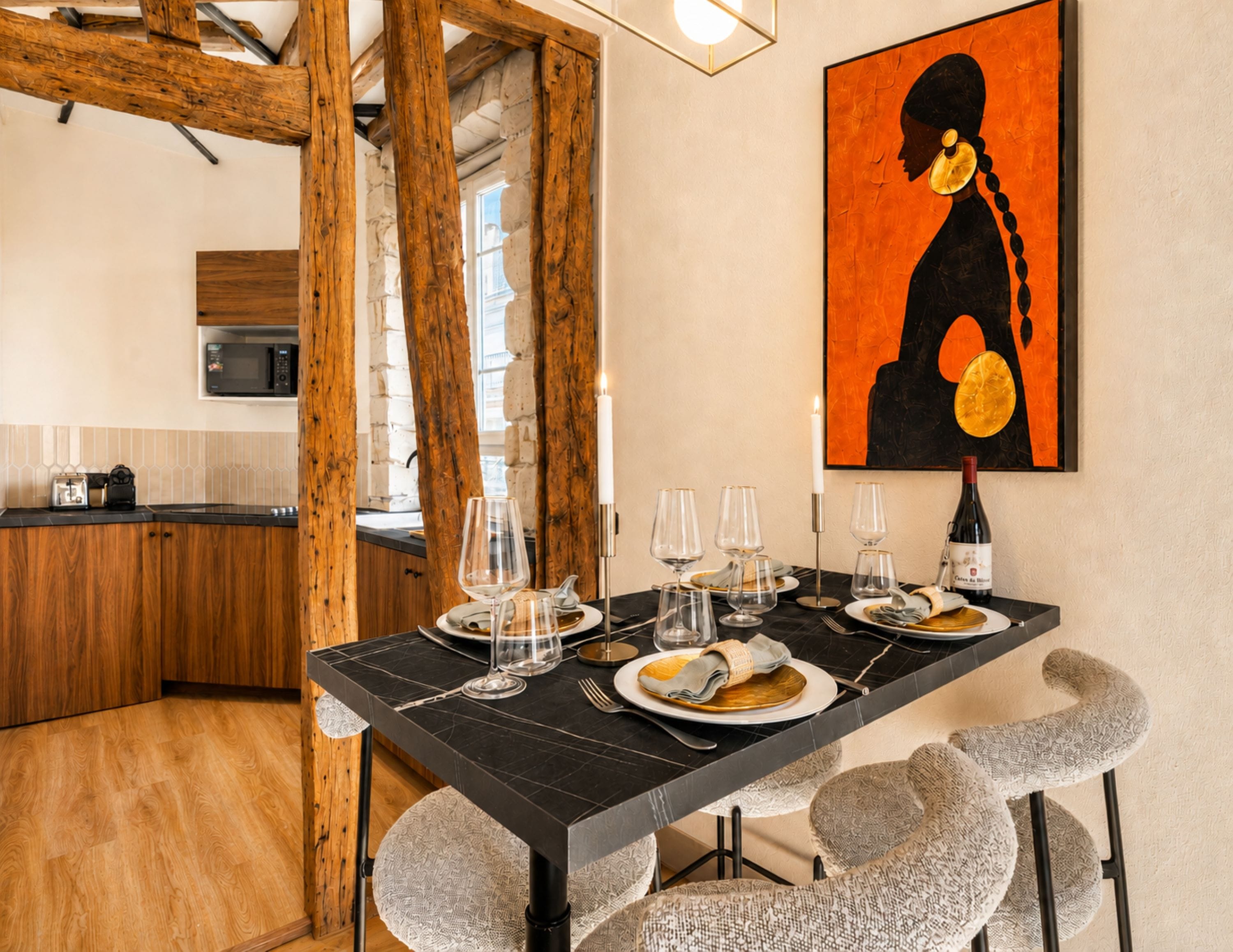 Un coin repas avec une table noire, des assiettes blanches et des verres à vin transparents. Une bougie est allumée sur la table, et un tableau est accroché au mur.
