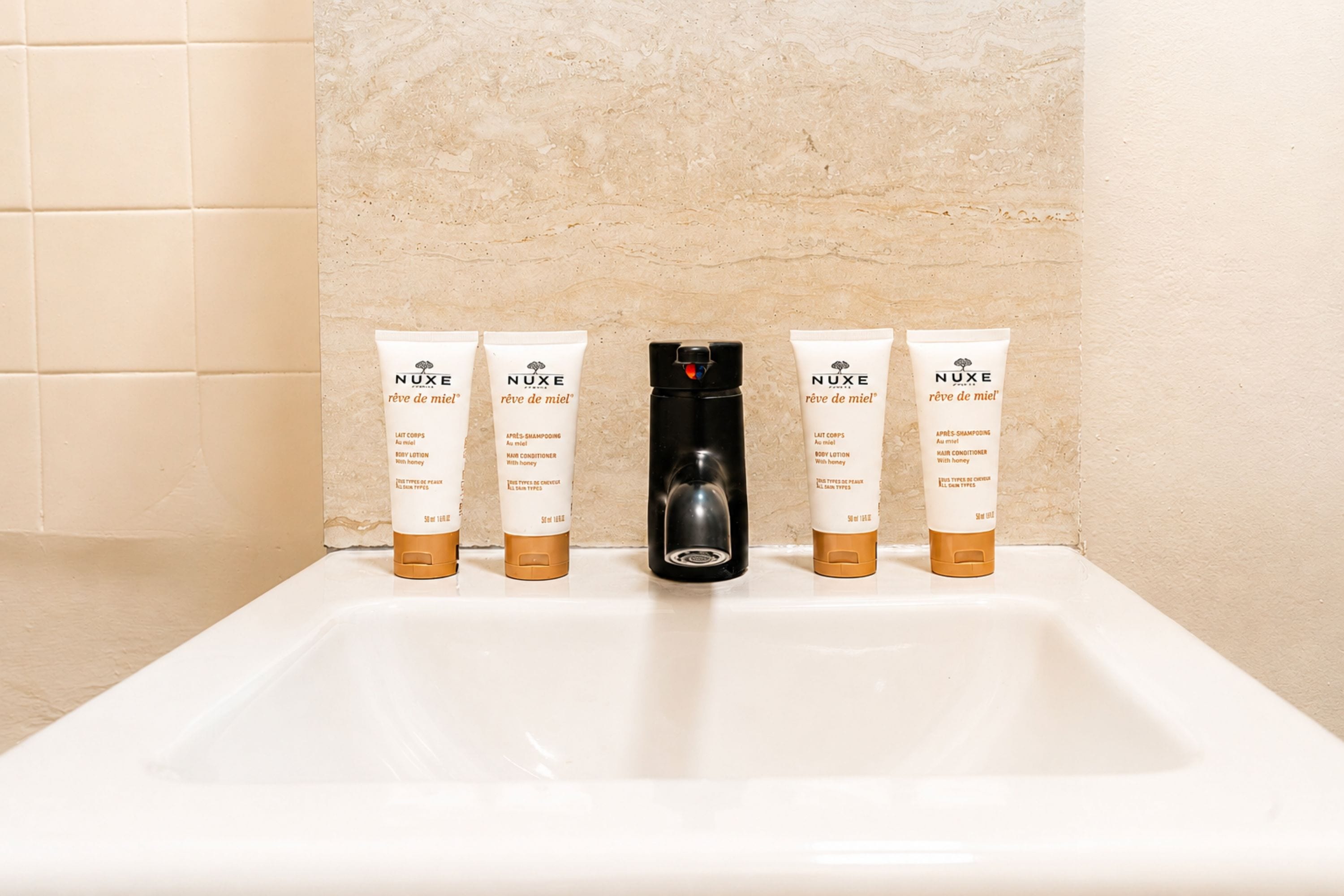A bathroom sink with four Nuxe body lotion and hair conditioner tubes and a black hairdryer.