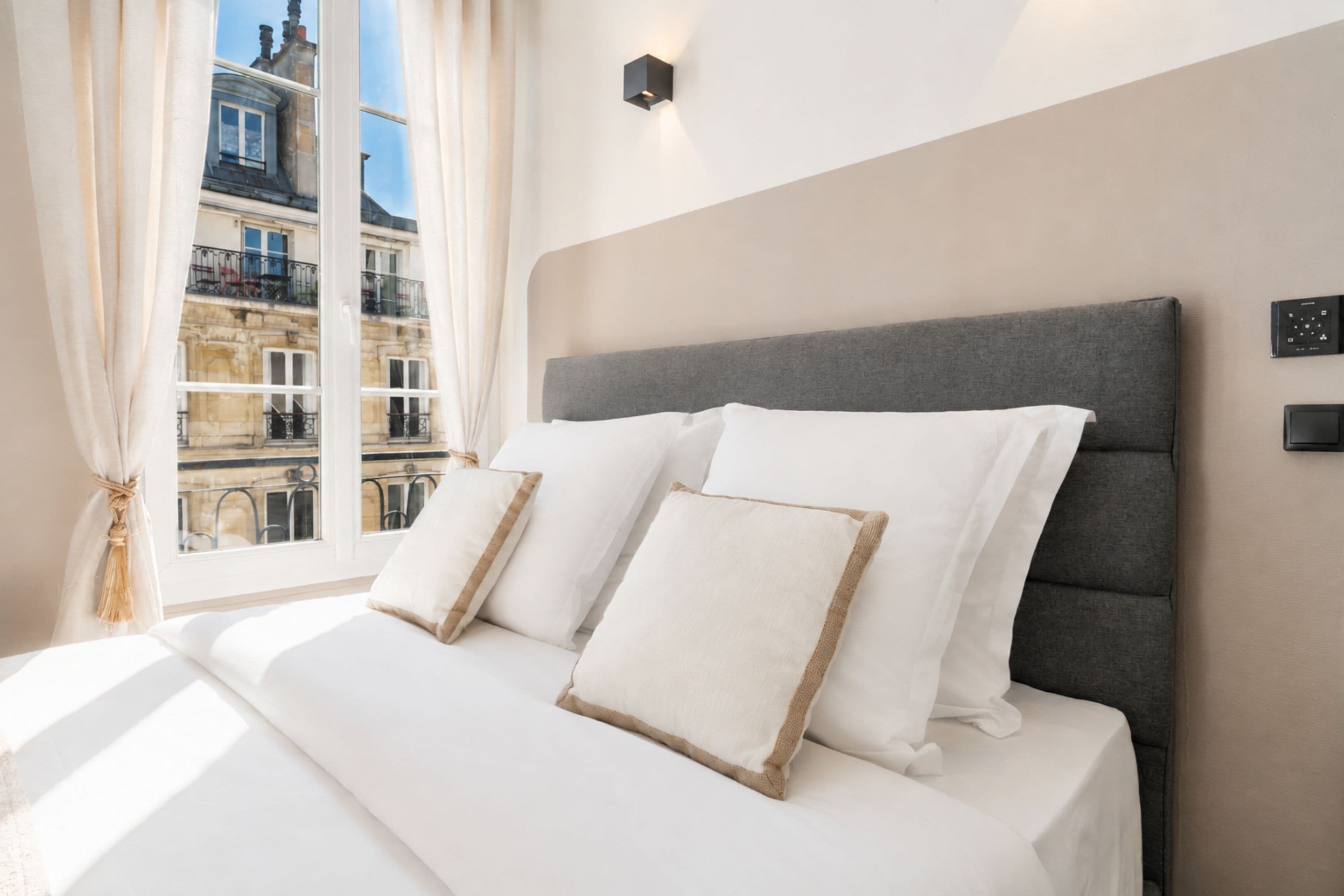 A hotel room with a large bed, a grey headboard, and a view of a building through the window.