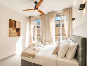 Un dormitorio con cama, cortinas, ventilador de techo, un cuadro y vistas a edificios a través de la ventana.