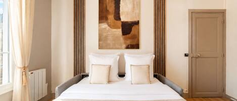 Une chambre d’hôtel avec un grand lit, deux oreillers, une tête de lit, une porte, un radiateur et une fenêtre avec des rideaux.