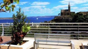 A bench overlooking a coastal view with a historic building and a clear sky. - Luxurious Gozo penthouse with sea view & pool (Għajnsielem)