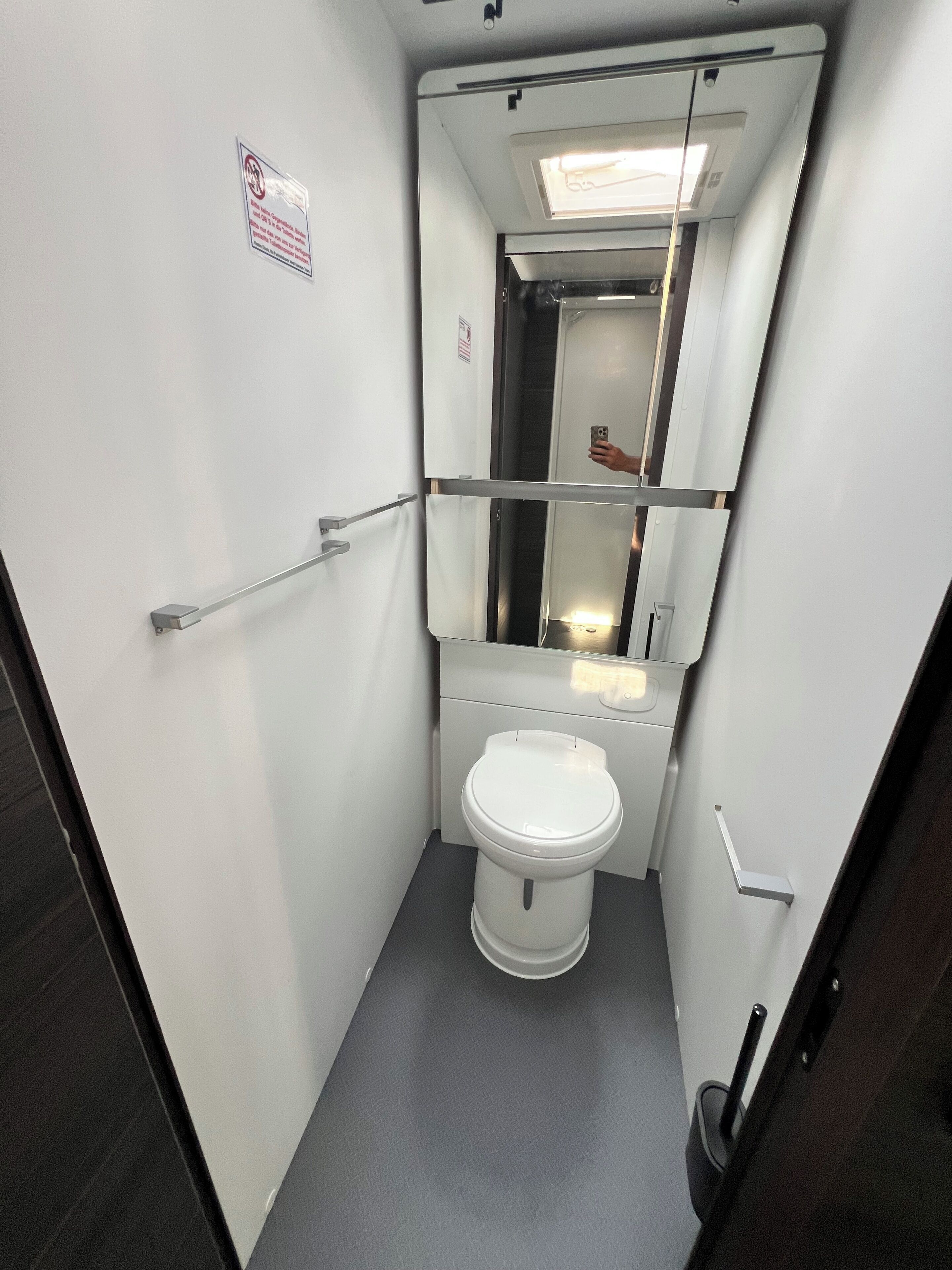 A compact, modern bathroom with a white toilet, a handrail, and a mirror.