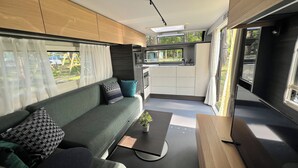 A modern RV interior with a sofa, a small table with a plant, a kitchenette, and a large window offering a view of the outdoors. - Luxury caravan 5 with whirlpool directly on the Achterwasser -holiday homes island Usedom (Lütow)