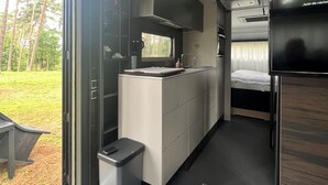 Una kitchenette compacta con una ventana, un pequeño contenedor de basura y una cama al fondo.